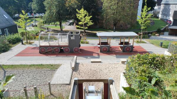 Der Mehrgenerationenspielplatz liegt im Ortskern von Kirchhundem hinter der Kirche. Ein Eisenbahn zum Spielen bietet auch Sitzmöglichkeiten im Schatten. Eine Rutsche führt den anliegenden Hang hinunter.