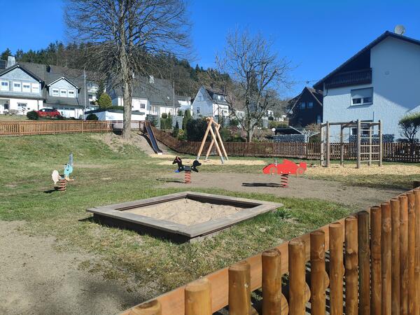 Ein eingezäunter Spielplatz mit verschiedenen Spielgeräten: In der Mitte stehen mehrere kleine Wippen, eine Schaukel, ein Klettergerüst und ein Sandkasten im Vordergrund. Am leicht abfallenden Hang rechts im Bild ist eine Rutsche integriert.