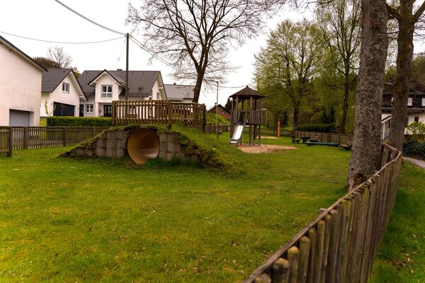 Der eingezäunte Spielplatz mitten im Wohngebiet ist von Bäumen beschatten und hat verschiedene Spielgeräute.